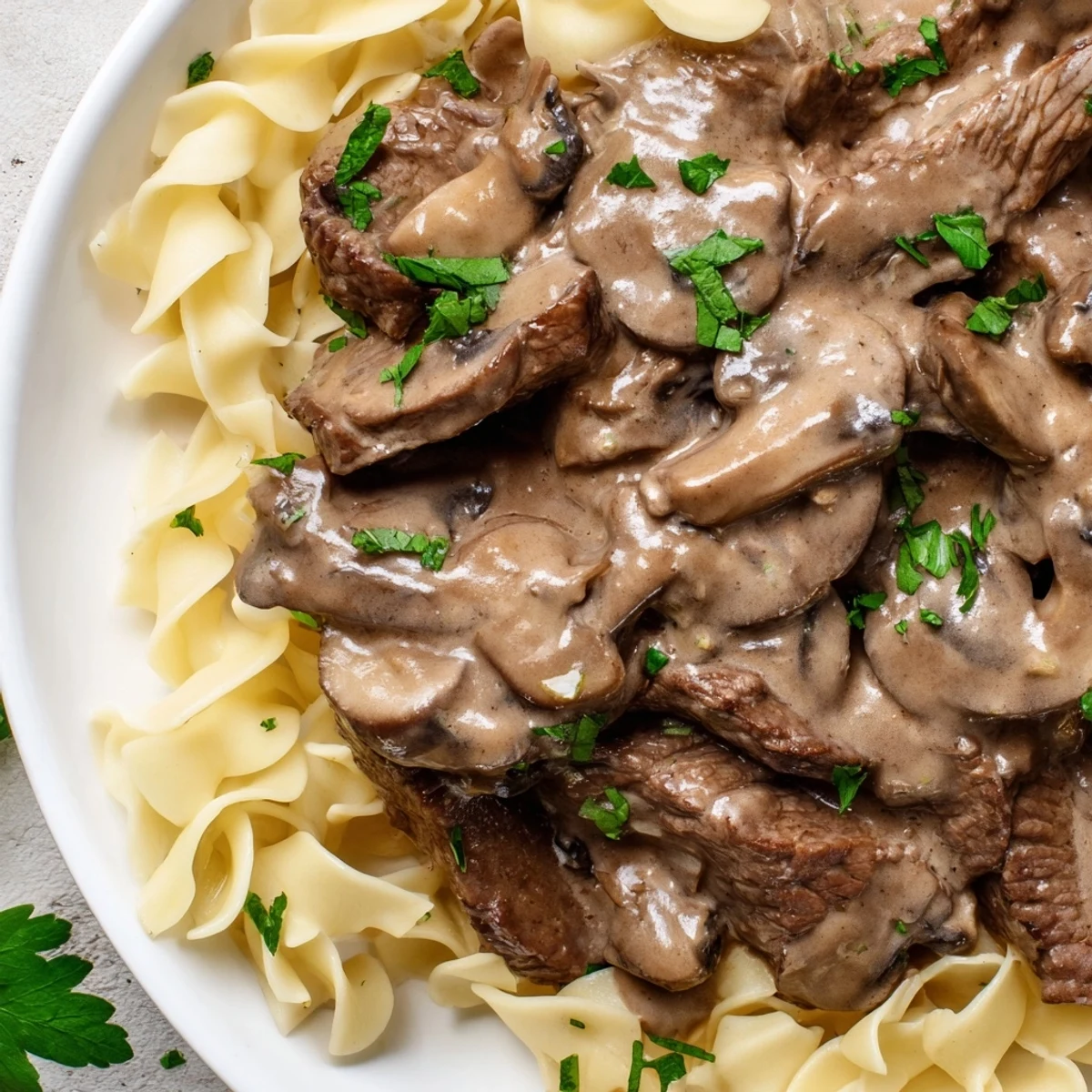 Steaming beef stroganoff with mushroom gravy is spooned over a bed of egg noodles on a white plate.