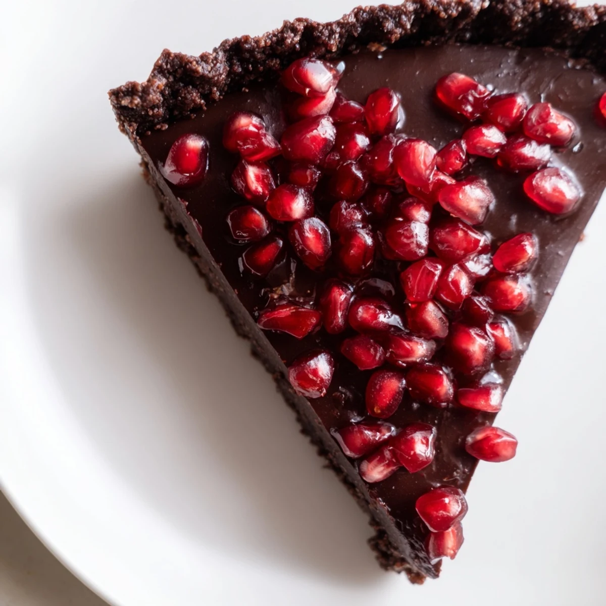 A glossy slice of Valentine Chocolate Pomegranate Tart showcases a rich chocolate ganache filling inside a dark cocoa crust.  