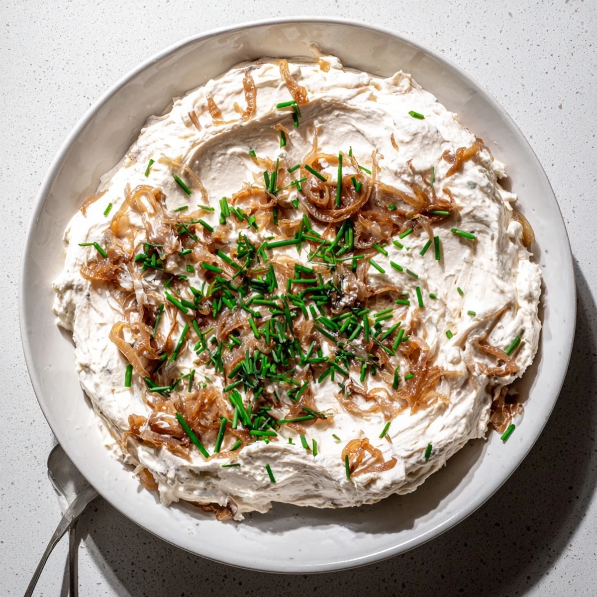 Creamy, golden-brown caramelized onion dip garnished with fresh chives, served in a rustic bowl beside crunchy potato chips.