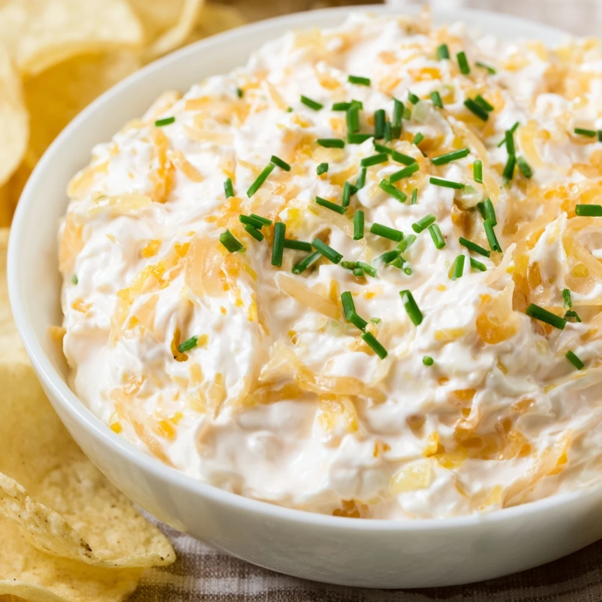 A rustic bowl of homemade Game Day Onion Dip surrounded by crunchy potato chips, garnished with fresh chives for a savory party snack.