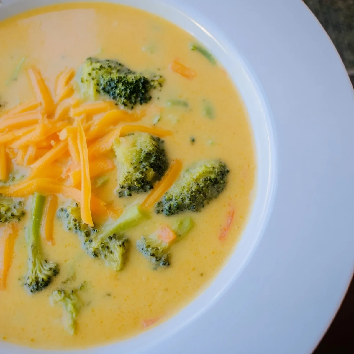 A steaming bowl of Creamy Broccoli Cheddar Soup garnished with extra cheese and a slice of crusty bread for dipping.  