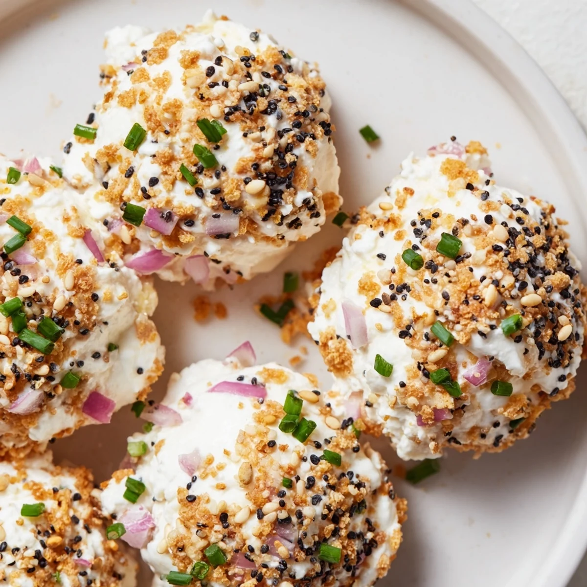 Golden-brown Everything Bagel Cottage Cheese Bites coated in sesame and poppy seeds rest on a white platter, garnished with fresh chives for a savory, gluten-free snack.