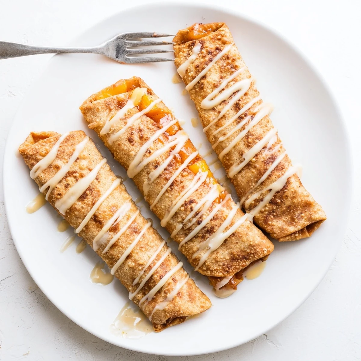 Golden-baked Peach Cobbler Egg Rolls glisten with vanilla glaze beside fresh peach slices on a rustic board.  