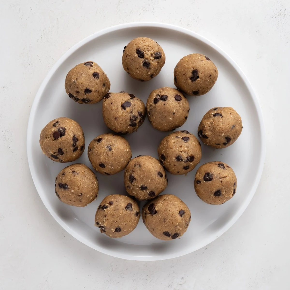 Bite-sized chocolate chip cookie dough protein balls rolled with mini dark chocolate chips on a white plate