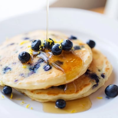 Fluffy pancakes with lemon zest and blueberries drizzled with warm maple syrup on a cozy plate.