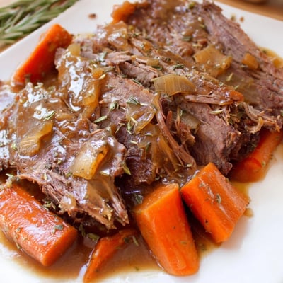 Golden-brown Slow Cooker Roast Beef with Gravy rests on a platter next to soft carrots, onions, and celery, with steam rising from the savory sauce.