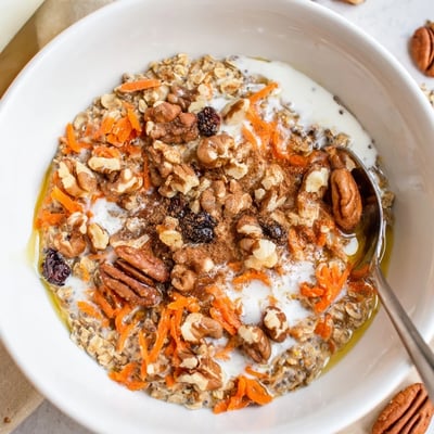 A spoon dips into the chilled Carrot Cake Overnight Oats breakfast, revealing layers of grated carrots and raisins.